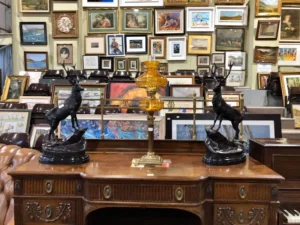 Antique wooden desk with two stag sculptures and an amber lamp, surrounded by framed paintings on the wall.