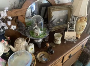 A wooden dresser displays vintage decor, including a tea set, framed art, a lamp, and various figurines and trinkets.