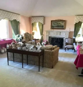 A traditional living room with vintage furniture, decorative glassware, and pink tablecloths covering multiple tables.
