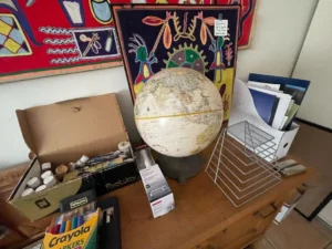 A globe, art supplies, and paperwork are arranged on a wooden table beneath colorful wall art.
