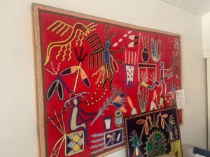Colorful, abstract wall art with figures, animals, and symbols on a red background, displayed in a room.