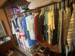 Clothes hanging on racks in a closet with shelves, boxes, and board games visible above and below.