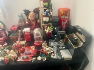 A table displays assorted Coca-Cola collectibles, holiday decorations, a plush moose, cameras, and various small items.