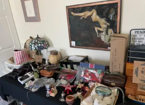 A table displays assorted vintage items, decor, and collectibles beneath a framed painting of a reclining figure on the wall.