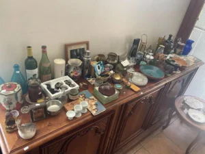 Assorted vintage items, dishes, and bottles are displayed on a wooden sideboard in a sunlit room.