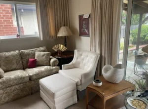 A beige armchair with an ottoman sits next to a floral sofa, wooden tables, and a large window with garden view.