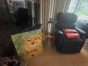 A painting of two cheetahs rests against a TV near a black recliner and a small wooden side table with items on it.