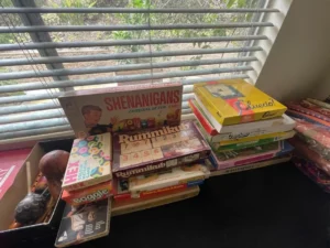 A stack of various board games, including Shenanigans, Rummikub, Clue, and Boggle, sits on a table by a window.