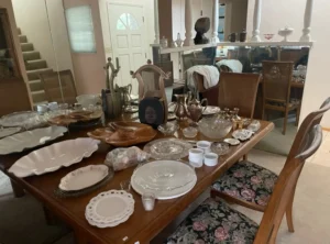 A wooden dining table displays assorted glassware, plates, and silverware, with floral-cushioned chairs around it.