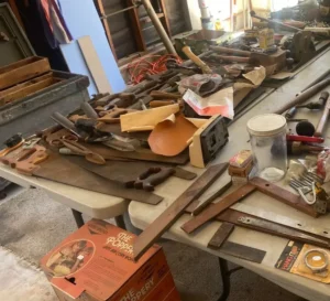 A variety of hand tools, sandpaper, and workshop items spread out on tables in a garage or workshop setting.