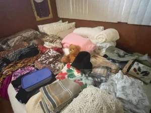 A bed covered with folded clothes, blankets, towels, a teddy bear, and various household items.