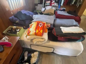Piles of folded clothes and blankets arranged on a bed, with boxes of miscellaneous items nearby in a cluttered room.