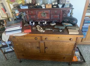 A wooden dresser and cabinet topped with books, jewelry, figurines, and various decorative items in a cluttered arrangement.